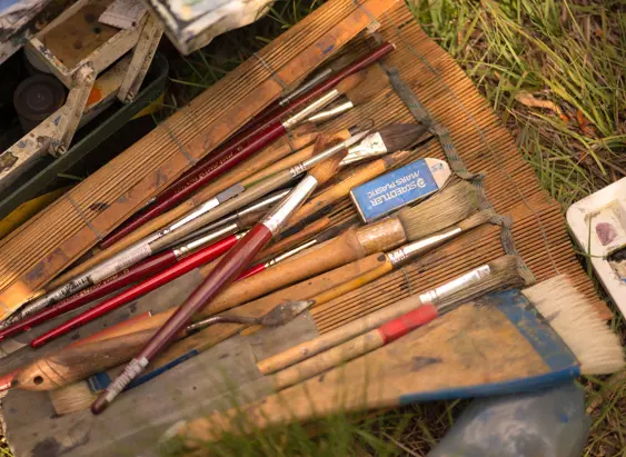 An artist's paintbrushes and palette laid out on a bamboo mat in the grass