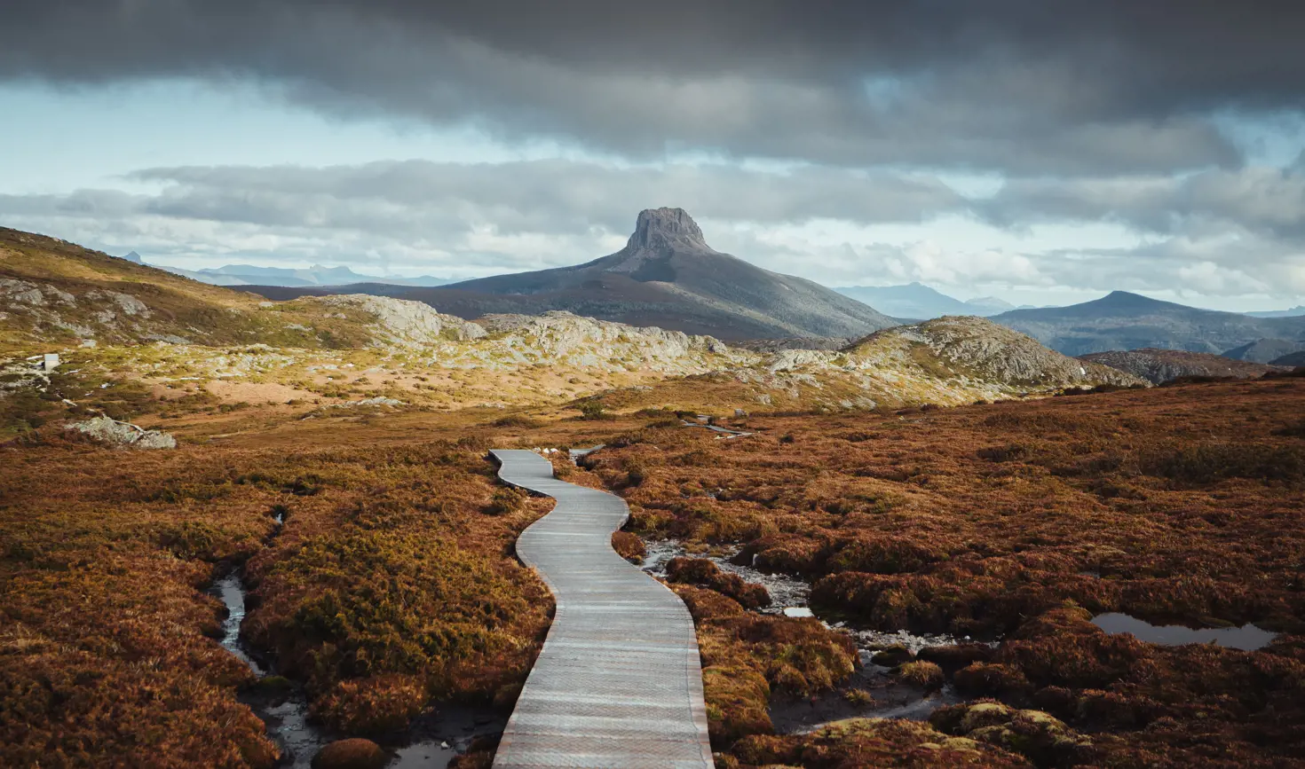 A boardwalk winds through alpine moorland toward a distinctive rocky peak on the Overland Track, Cradle Mountain