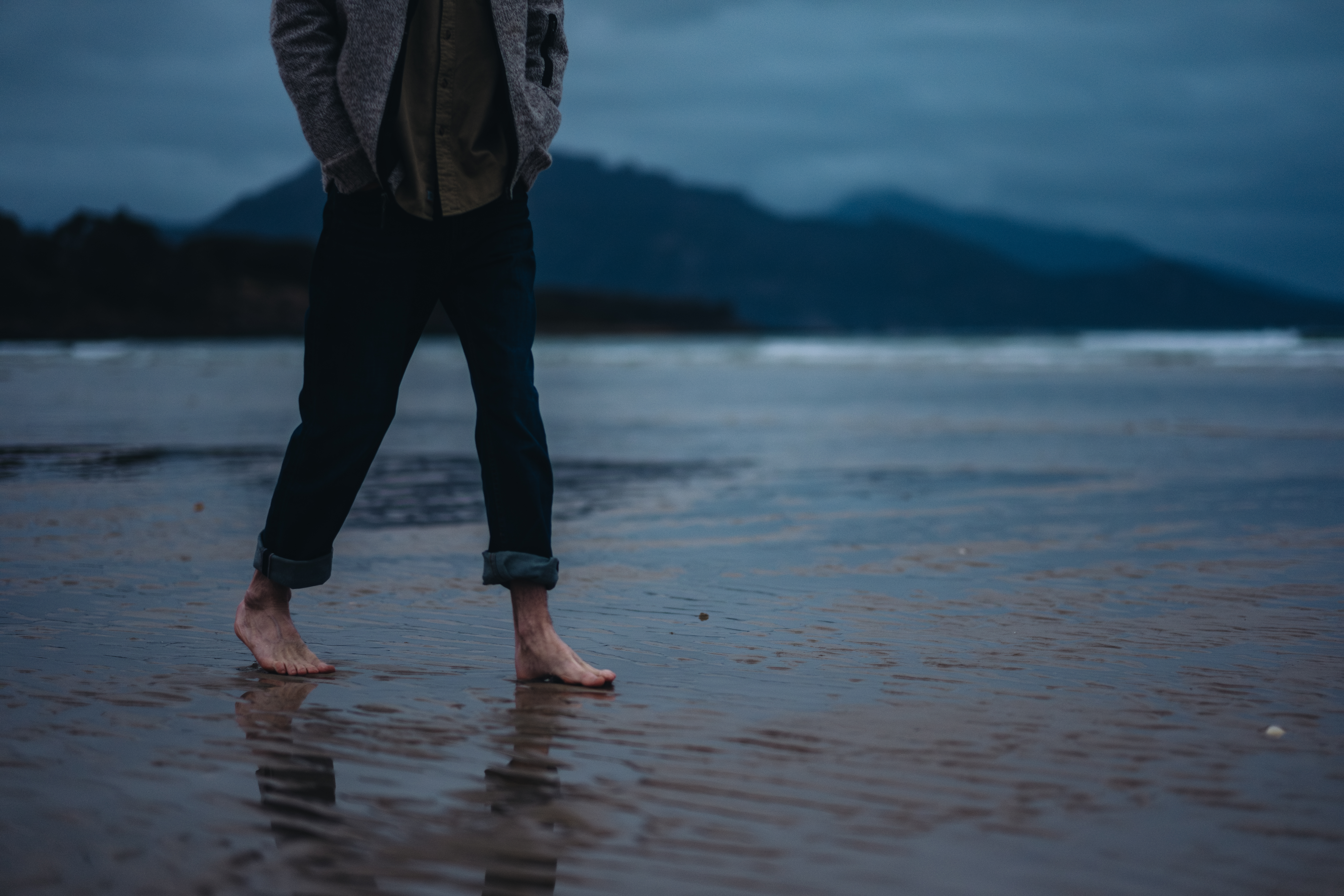 A person walks barefoot across wet sand on Sandpiper Beach with mountains and moody blue skies in the background