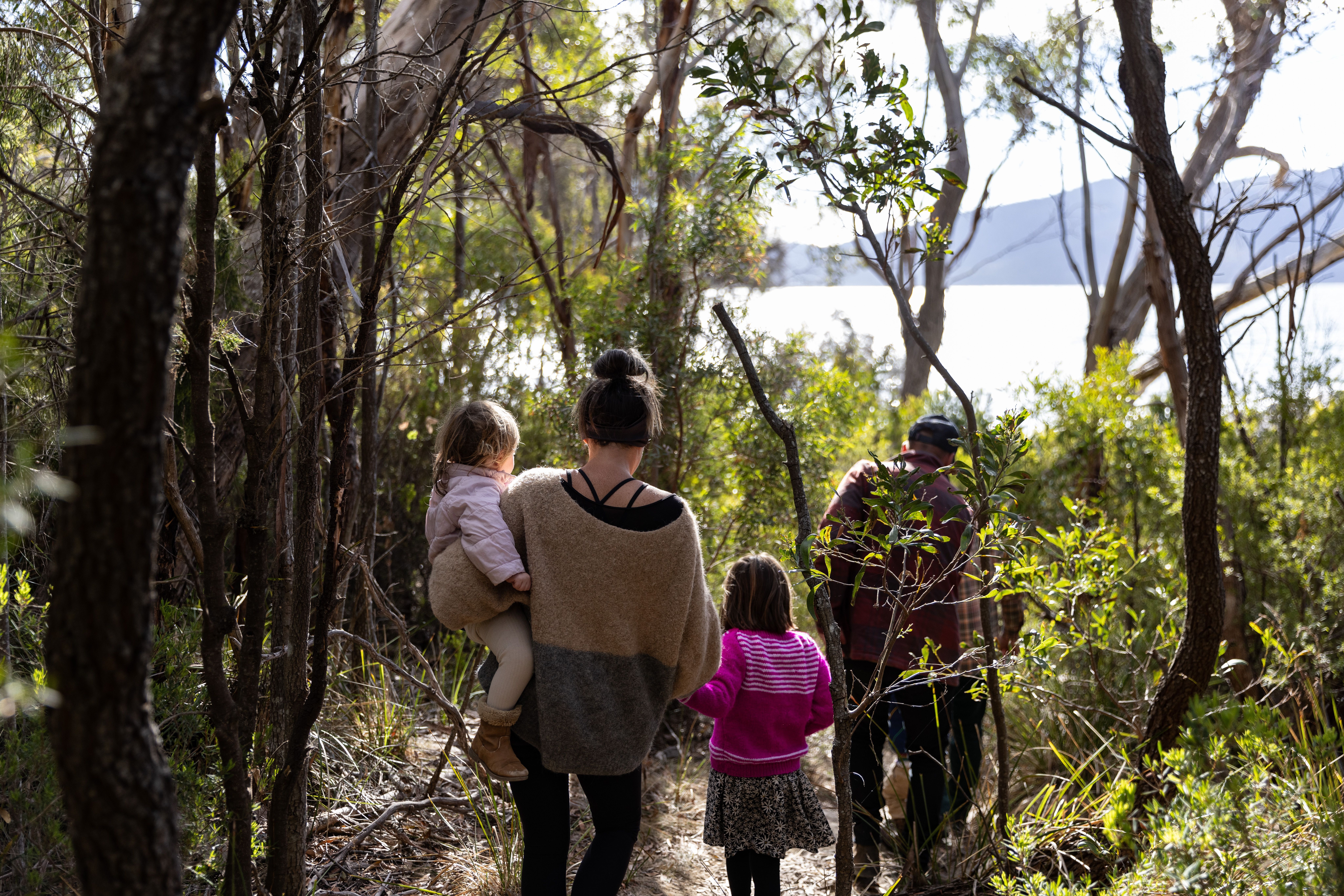 A family walks along a narrow bush track towards the water at Mandala on Bruny Island, with a woman carrying a young child