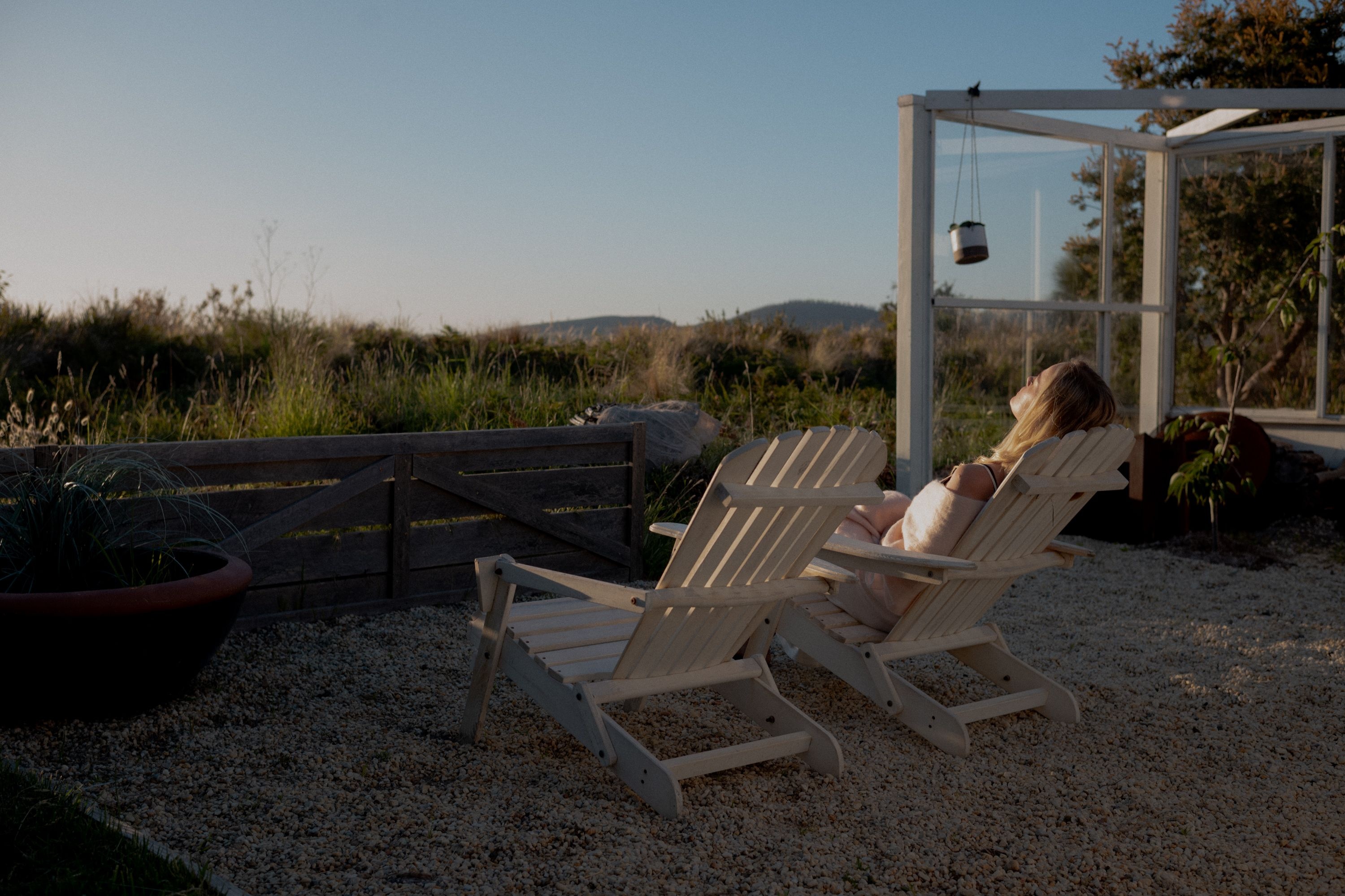 A person reclines on a wooden outdoor chair in warm light with rural views and a glass structure at Dunalley Bay Sauna