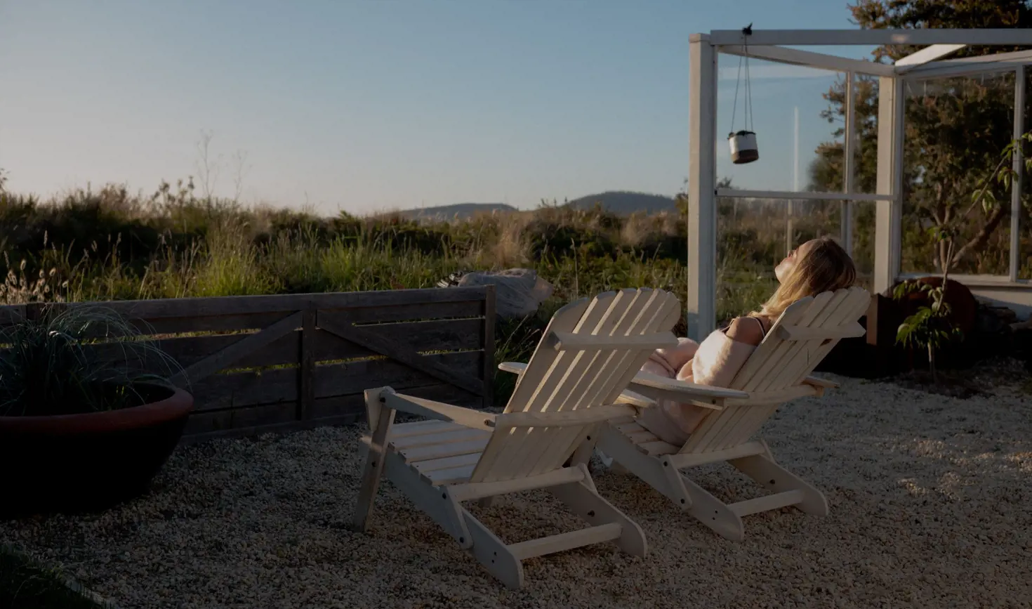 A person reclines on a wooden outdoor chair in warm light with rural views and a glass structure at Dunalley Bay Sauna