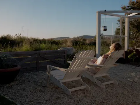 A person reclines on a wooden outdoor chair in warm light with rural views and a glass structure at Dunalley Bay Sauna