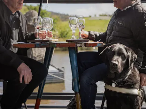 Two people enjoy a wine tasting flight at an outdoor table at Puddleduck Vineyard in Richmond accompanied by a guide dog
