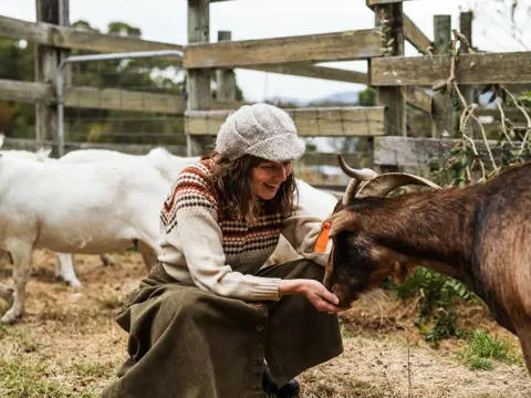 A smiling woman in a knitted hat crouches to feed a brown goat from her hand at Lavender Goat Farm in Petcheys Bay