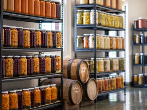 Shelves of preserved fruit and pickles in glass jars alongside wooden barrels at The Agrarian Kitchen in New Norfolk