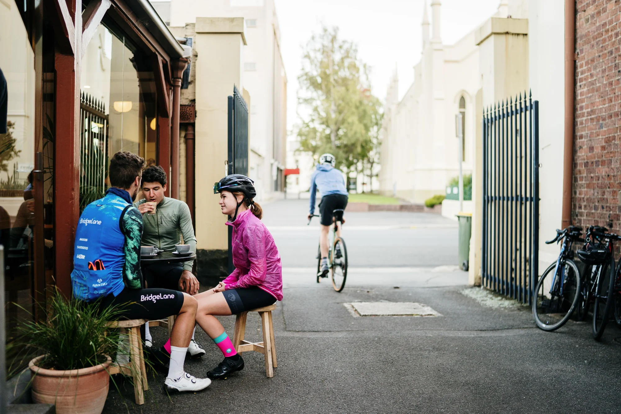 Three cyclists in riding gear sit outside Valley Coffee on stools while another cyclist rides past in a laneway, Launceston