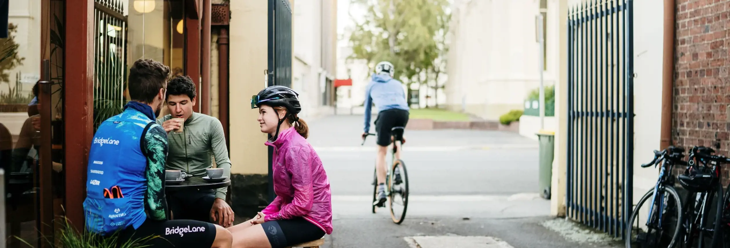 Three cyclists in riding gear sit outside Valley Coffee on stools while another cyclist rides past in a laneway, Launceston
