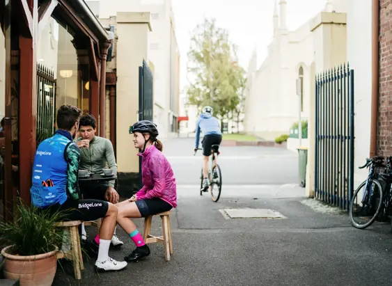 Three cyclists in riding gear sit outside Valley Coffee on stools while another cyclist rides past in a laneway, Launceston