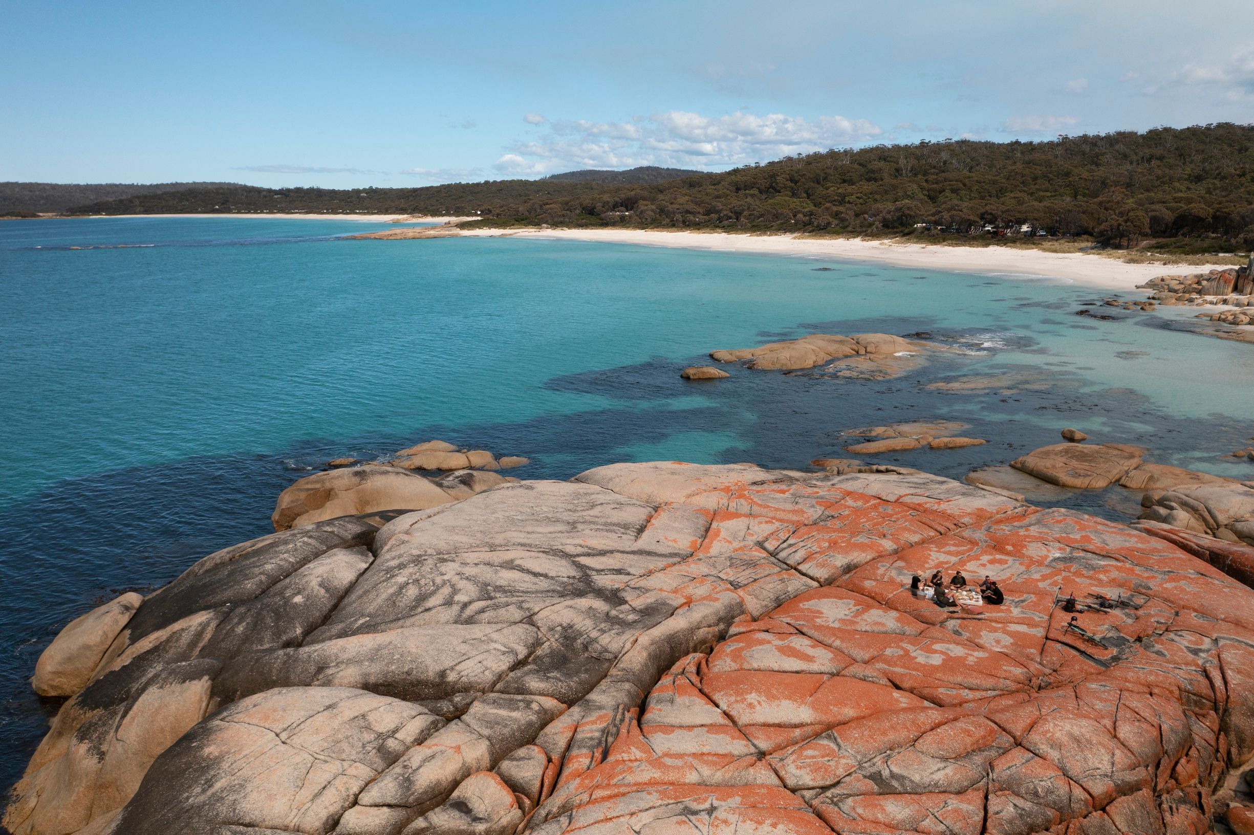 A small group has a picnic on orange lichen-covered granite rocks overlooking turquoise water and white beach on the east coast