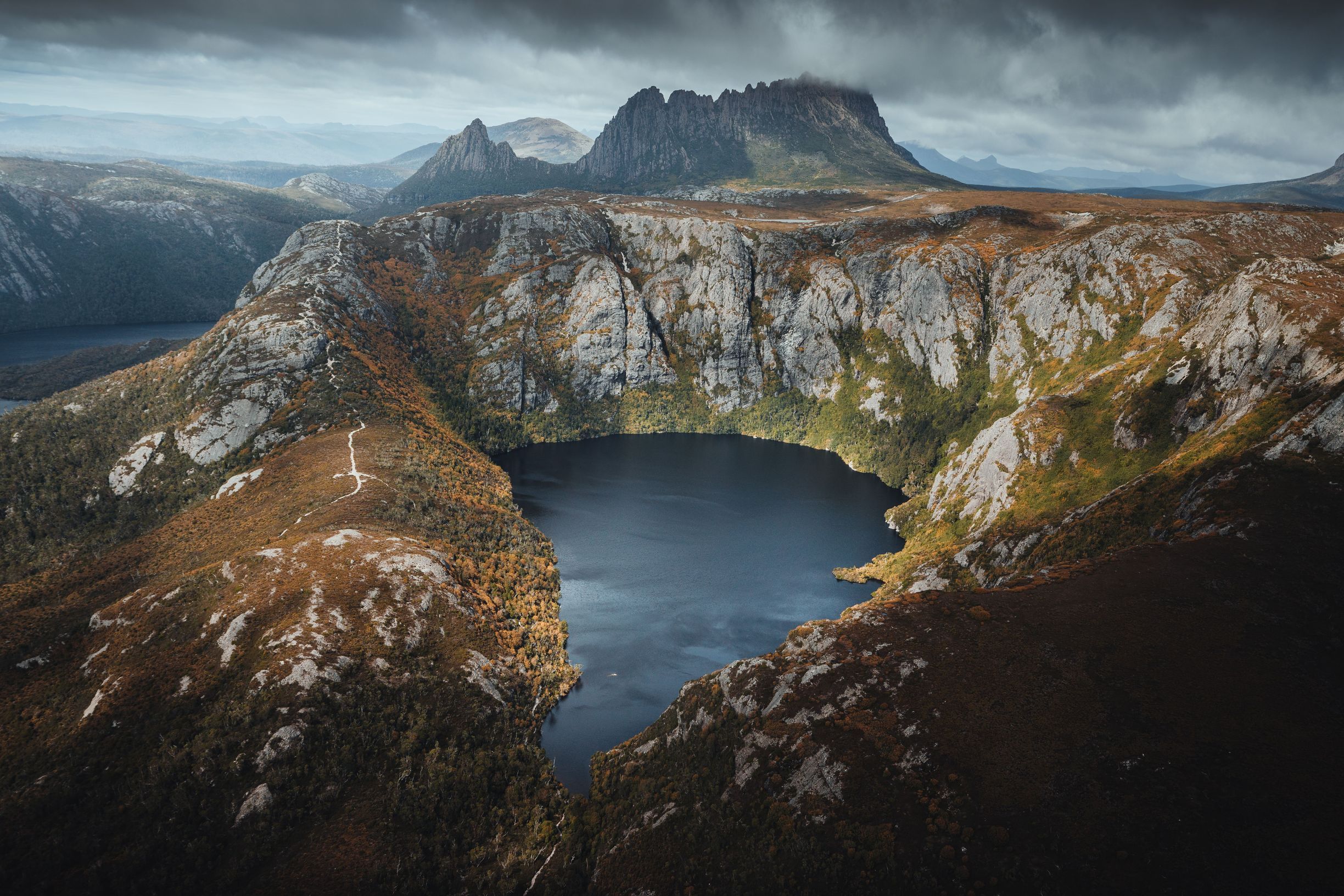Aerial view of Crater Lake nestled between steep rocky cliffs with the jagged peaks of Cradle Mountain under moody skies