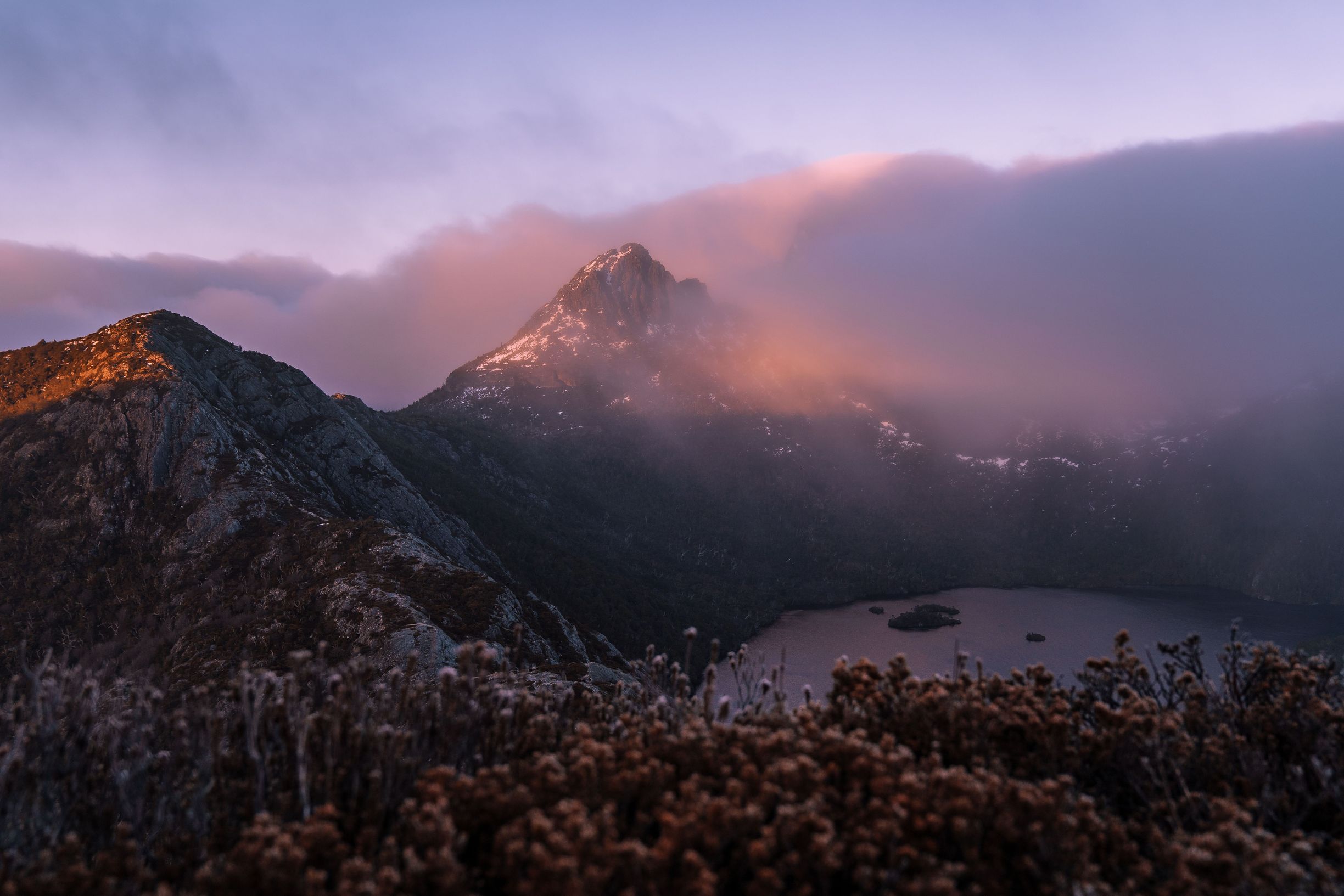 Cradle Mountain peak emerges from pink and purple clouds with Dove Lake below viewed from Mount Campbell