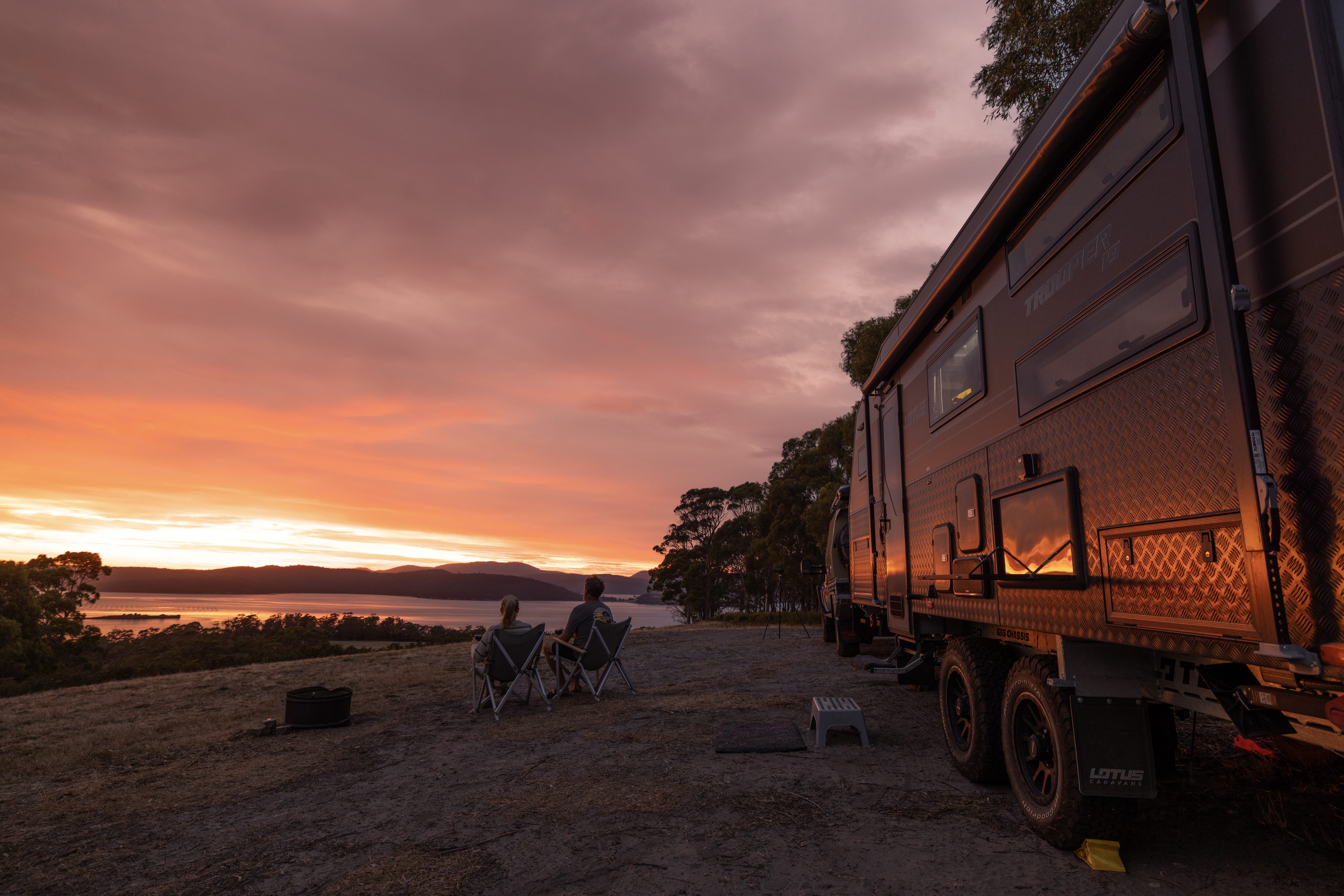 Two people in camp chairs watch the sunset over the water beside a caravan at East Beach Tourist Park, Low Head