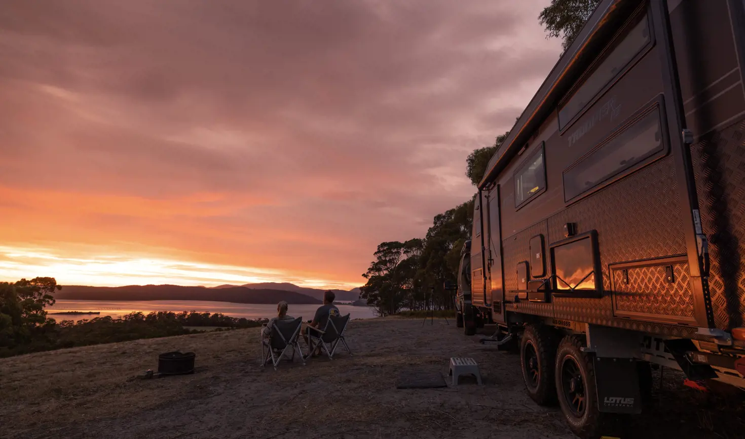 Two people in camp chairs watch the sunset over the water beside a caravan at East Beach Tourist Park, Low Head