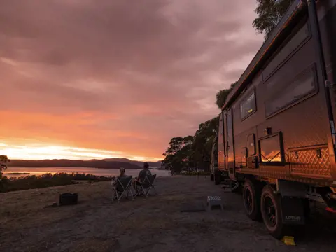 Two people in camp chairs watch the sunset over the water beside a caravan at East Beach Tourist Park, Low Head