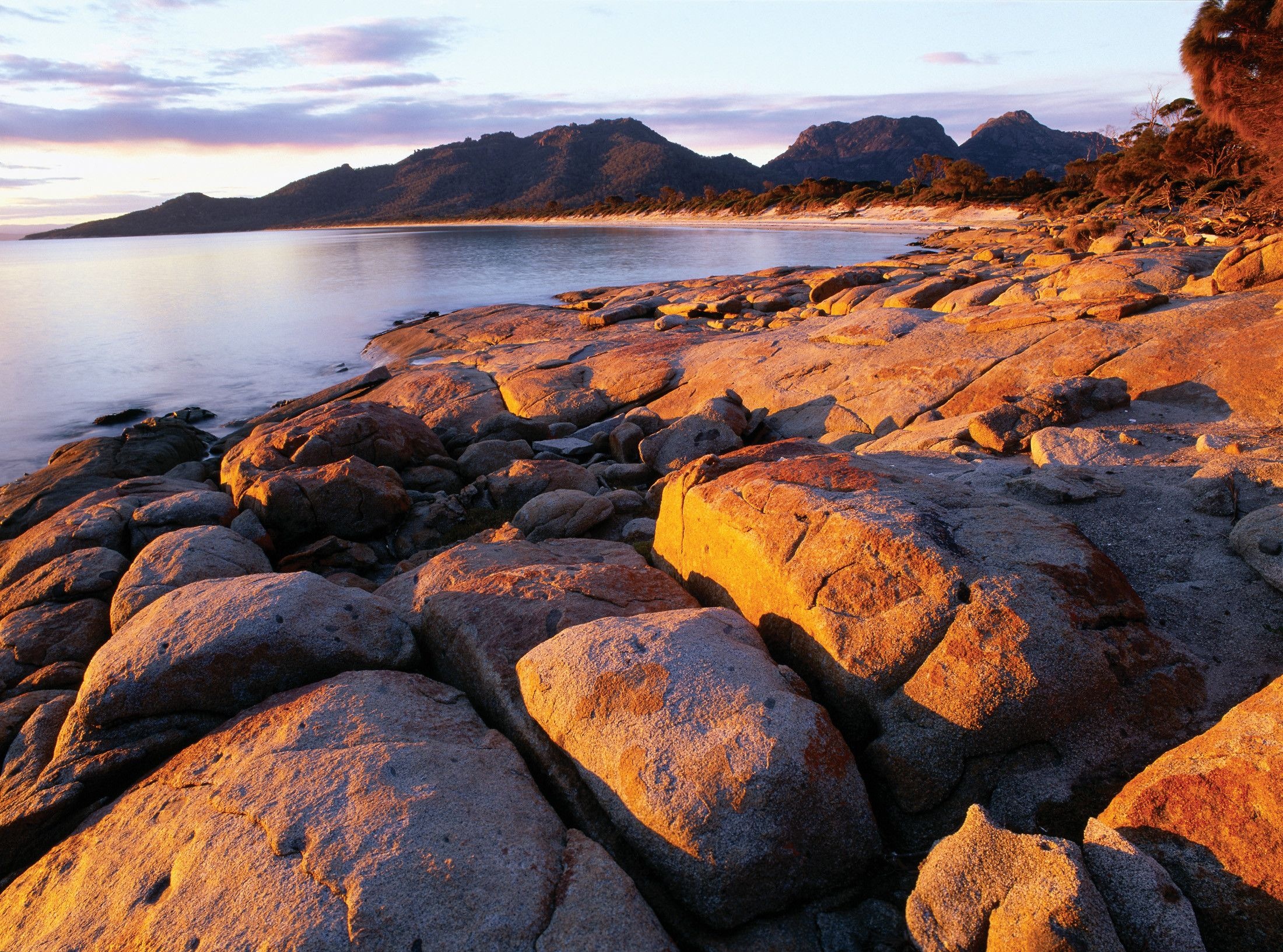 Granite boulders glow in warm light along the shore of Hazards Beach with The Hazards mountains beyond, Freycinet National Park