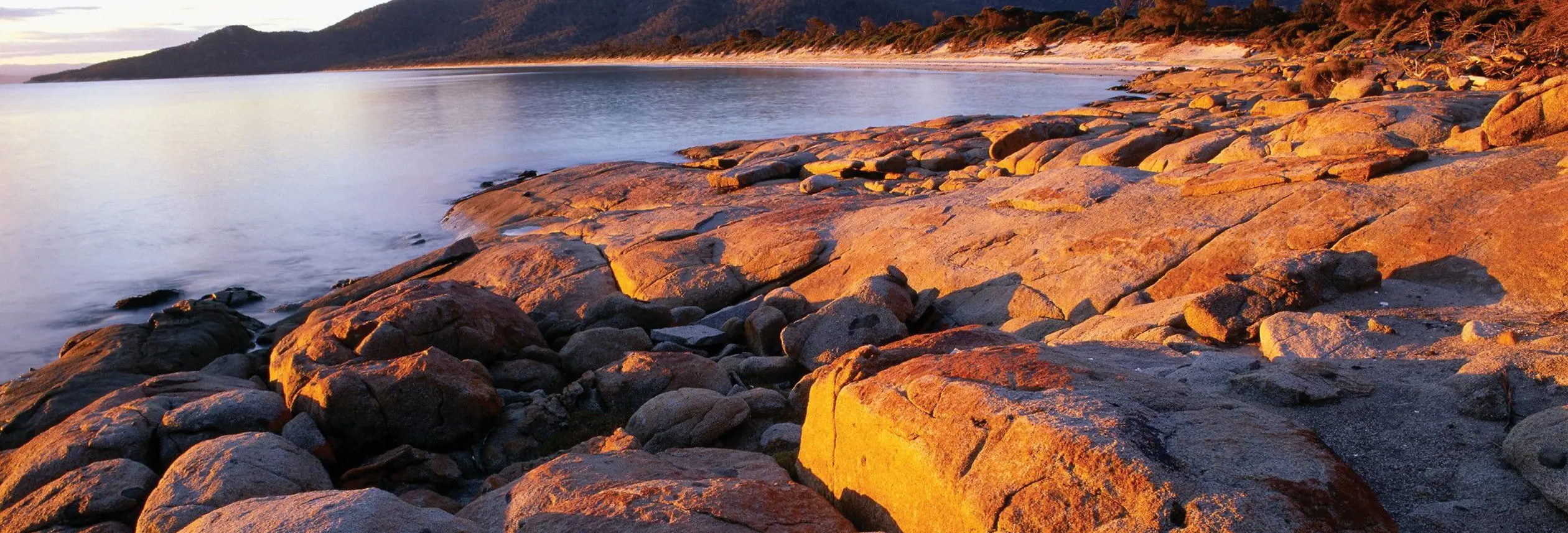 Granite boulders glow in warm light along the shore of Hazards Beach with The Hazards mountains beyond, Freycinet National Park