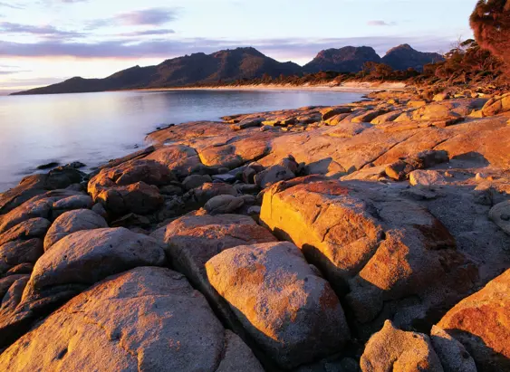 Granite boulders glow in warm light along the shore of Hazards Beach with The Hazards mountains beyond, Freycinet National Park