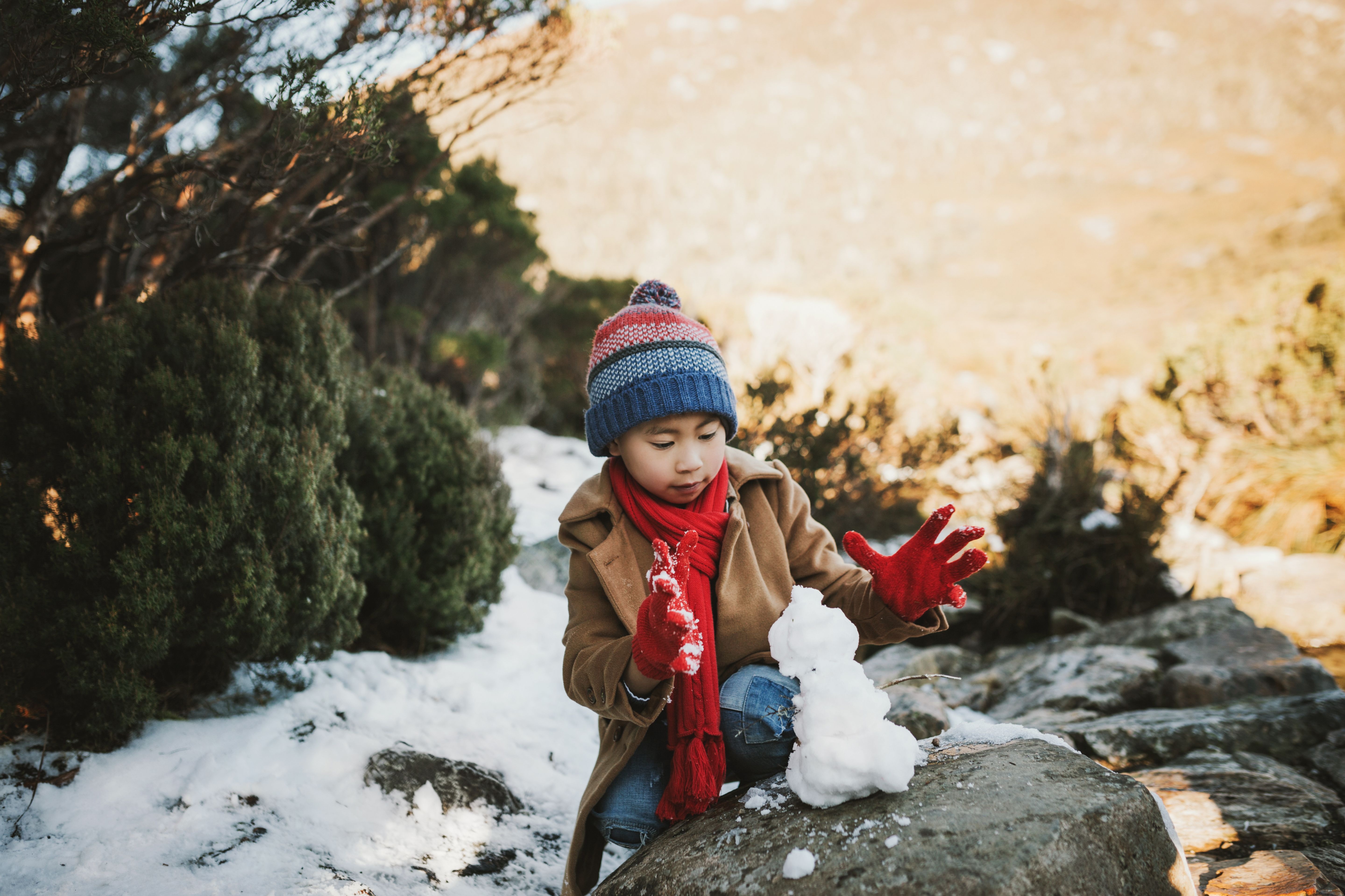 A young child in a beanie and red scarf builds a small snowman on a rock at Cradle Mountain