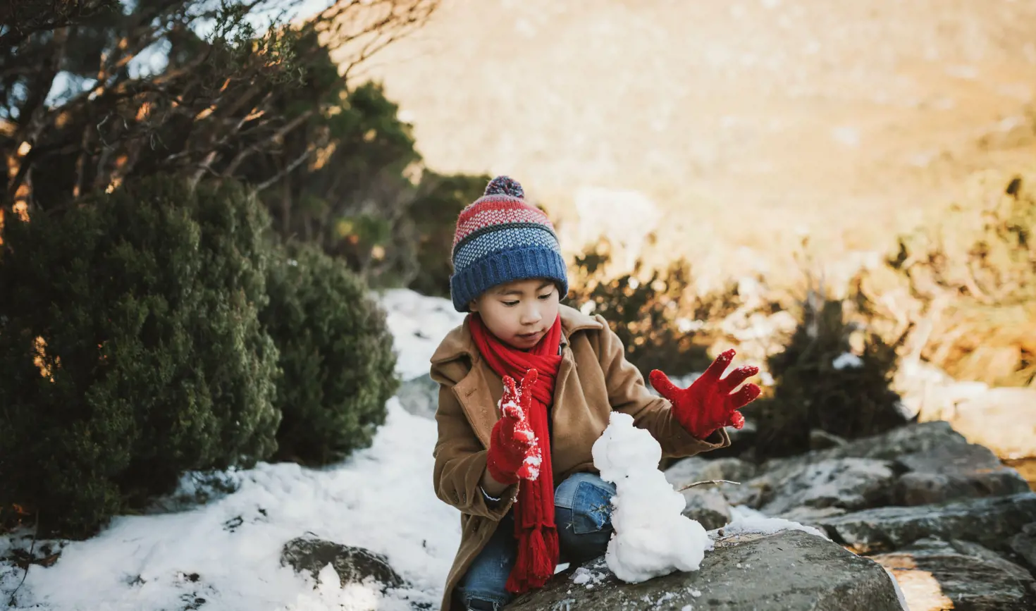 A young child in a beanie and red scarf builds a small snowman on a rock at Cradle Mountain