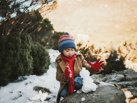 A young child in a beanie and red scarf builds a small snowman on a rock at Cradle Mountain