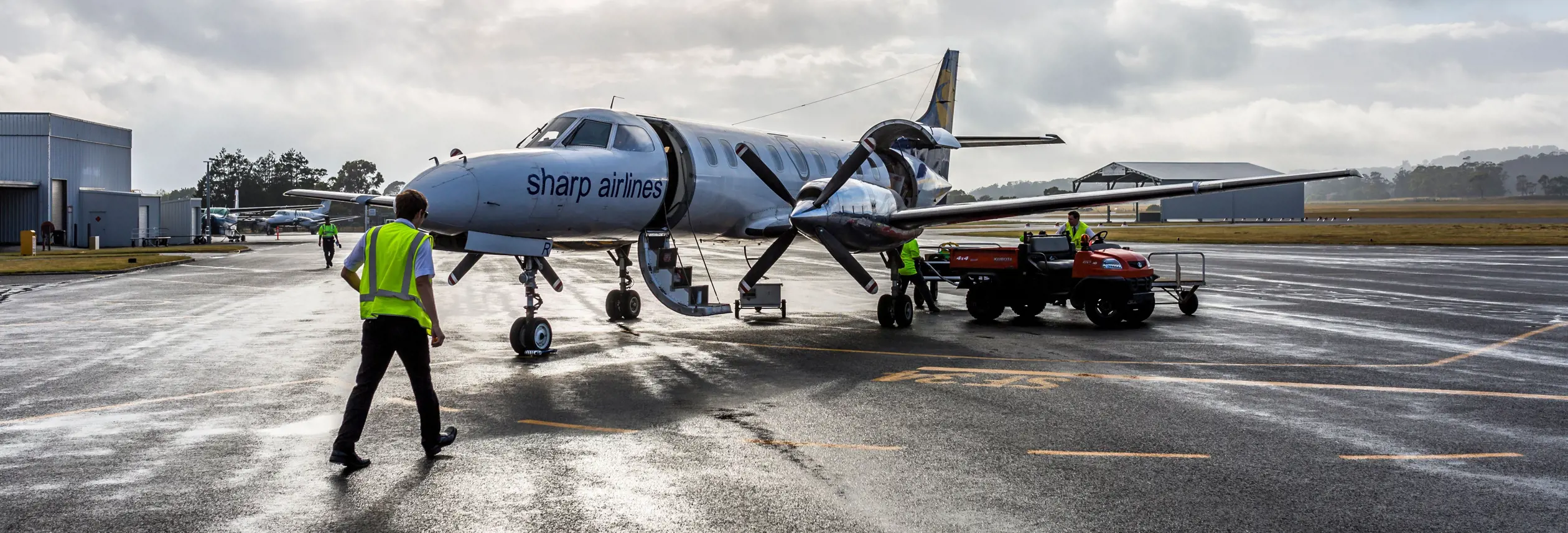 Ground crew in high visibility vests working around a Sharp Airlines propeller aircraft on a wet tarmac, King Island Airport