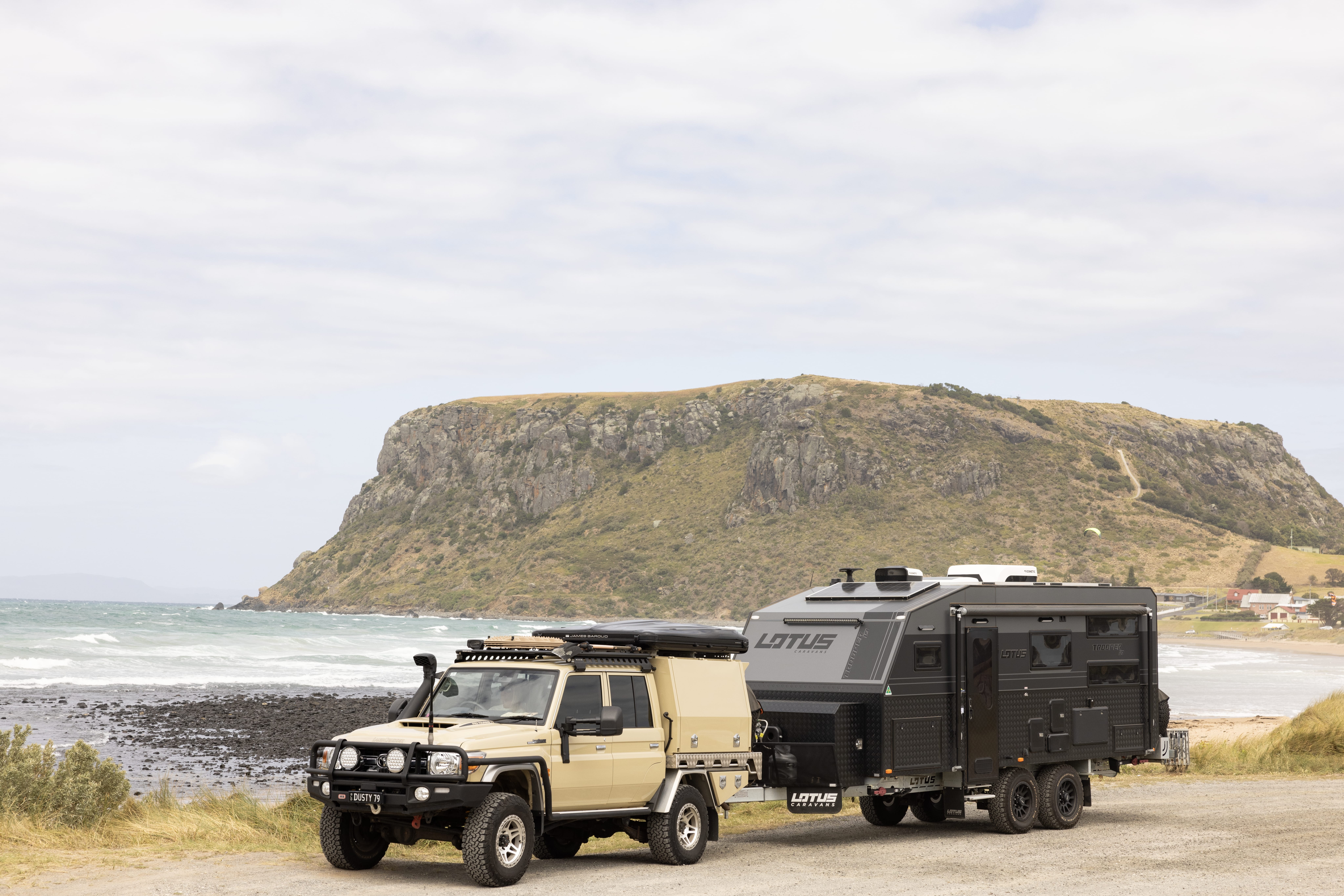 A 4WD towing a Lotus caravan parked on the foreshore with The Nut and the beach beyond, Stanley