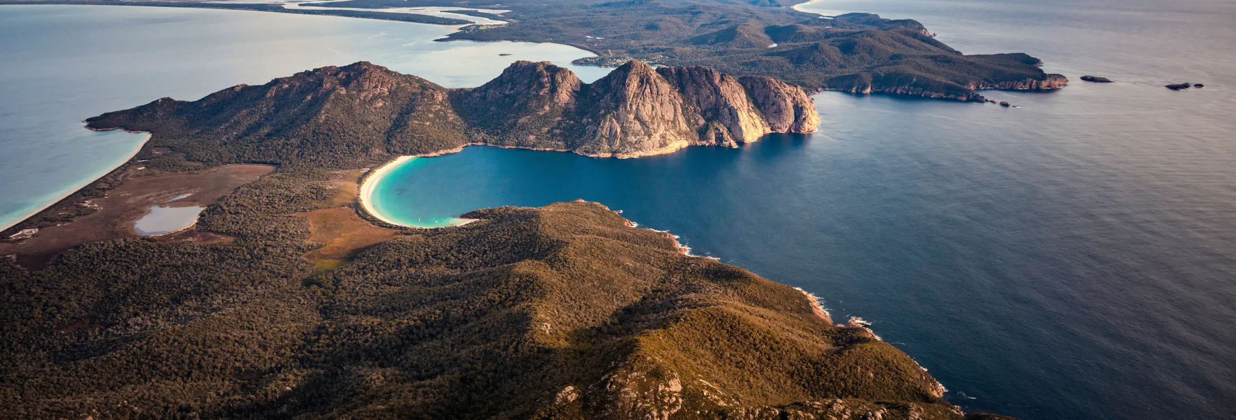 Aerial view of Wineglass Bay's crescent beach and turquoise water flanked by coastal cliffs, Freycinet National Park
