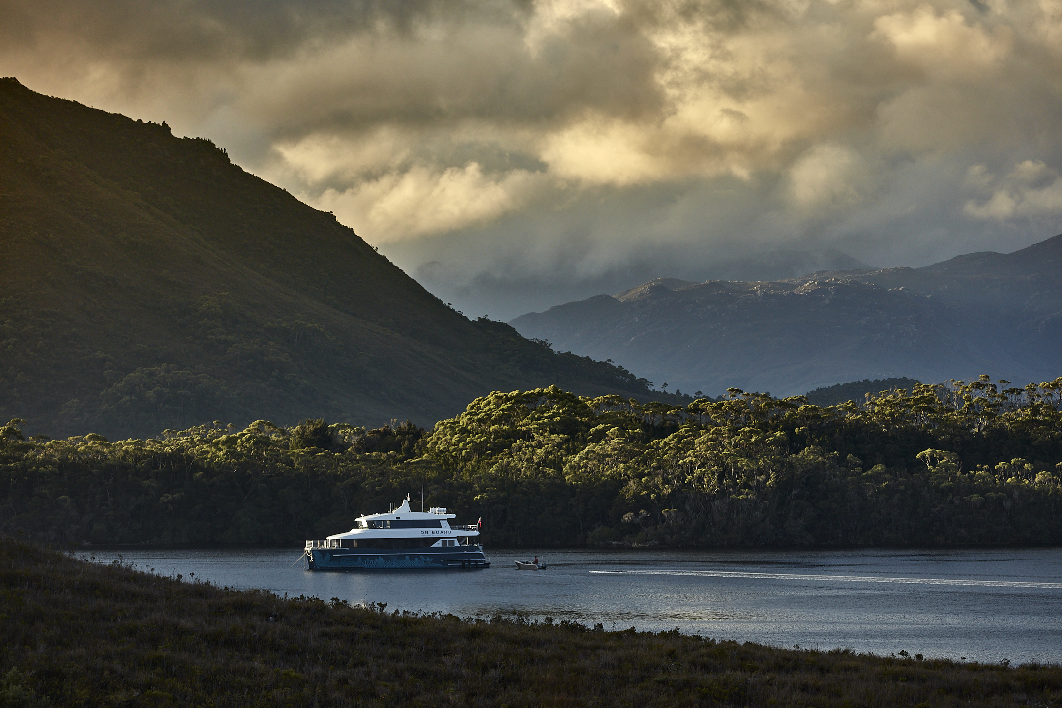 The On Board cruise vessel on calm water in Bathurst Harbour with forested mountains beyond, Southwest National Park