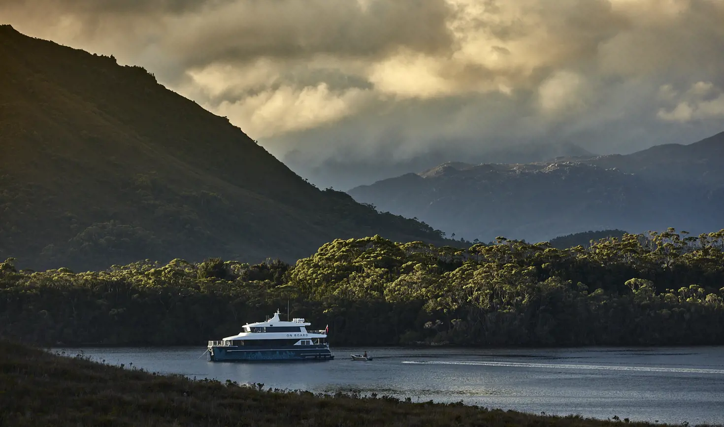 The On Board cruise vessel on calm water in Bathurst Harbour with forested mountains beyond, Southwest National Park