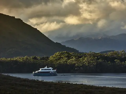 The On Board cruise vessel on calm water in Bathurst Harbour with forested mountains beyond, Southwest National Park