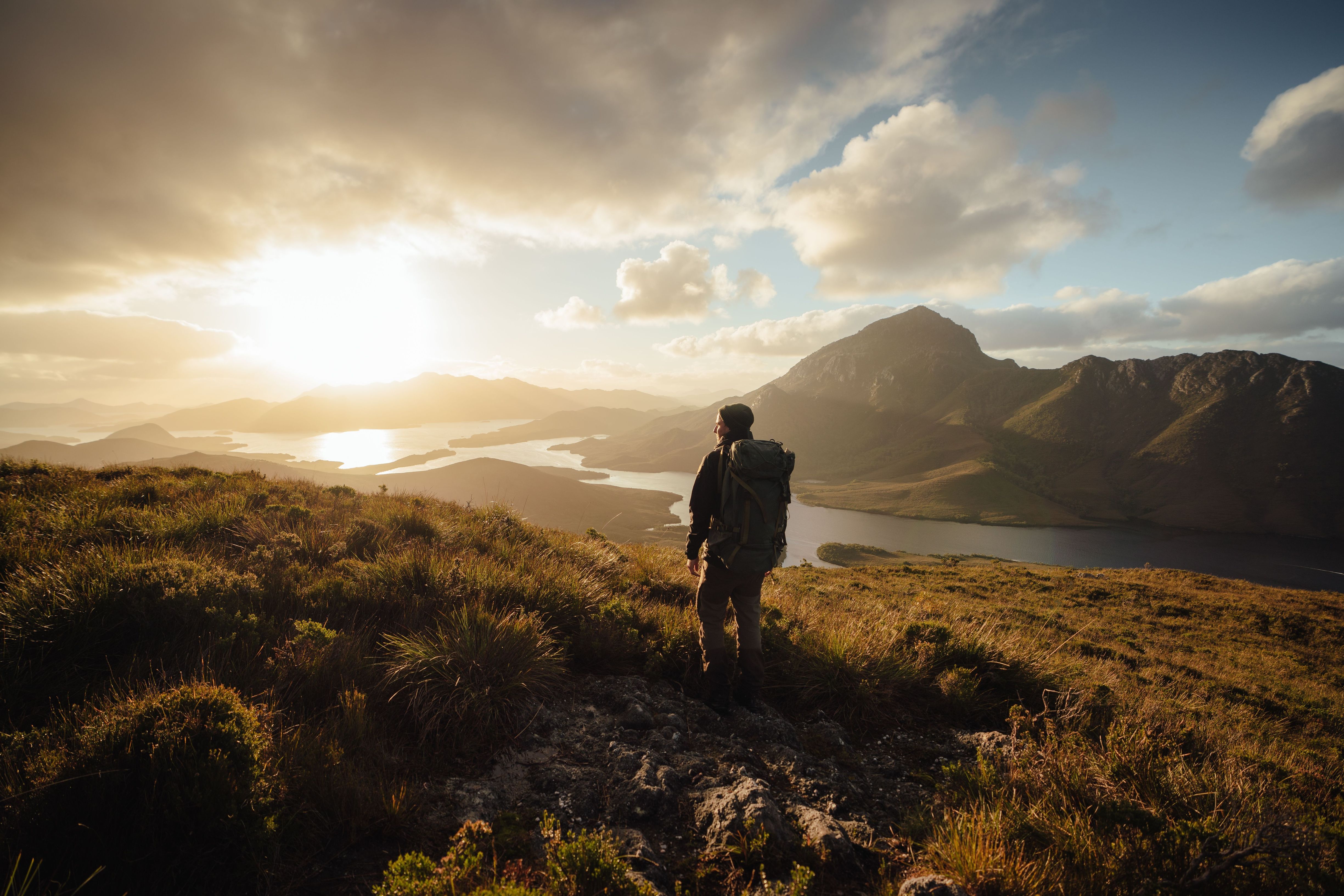 Hiker with a backpack on a grassy ridge looking out over the water and mountains at Bathurst Harbour