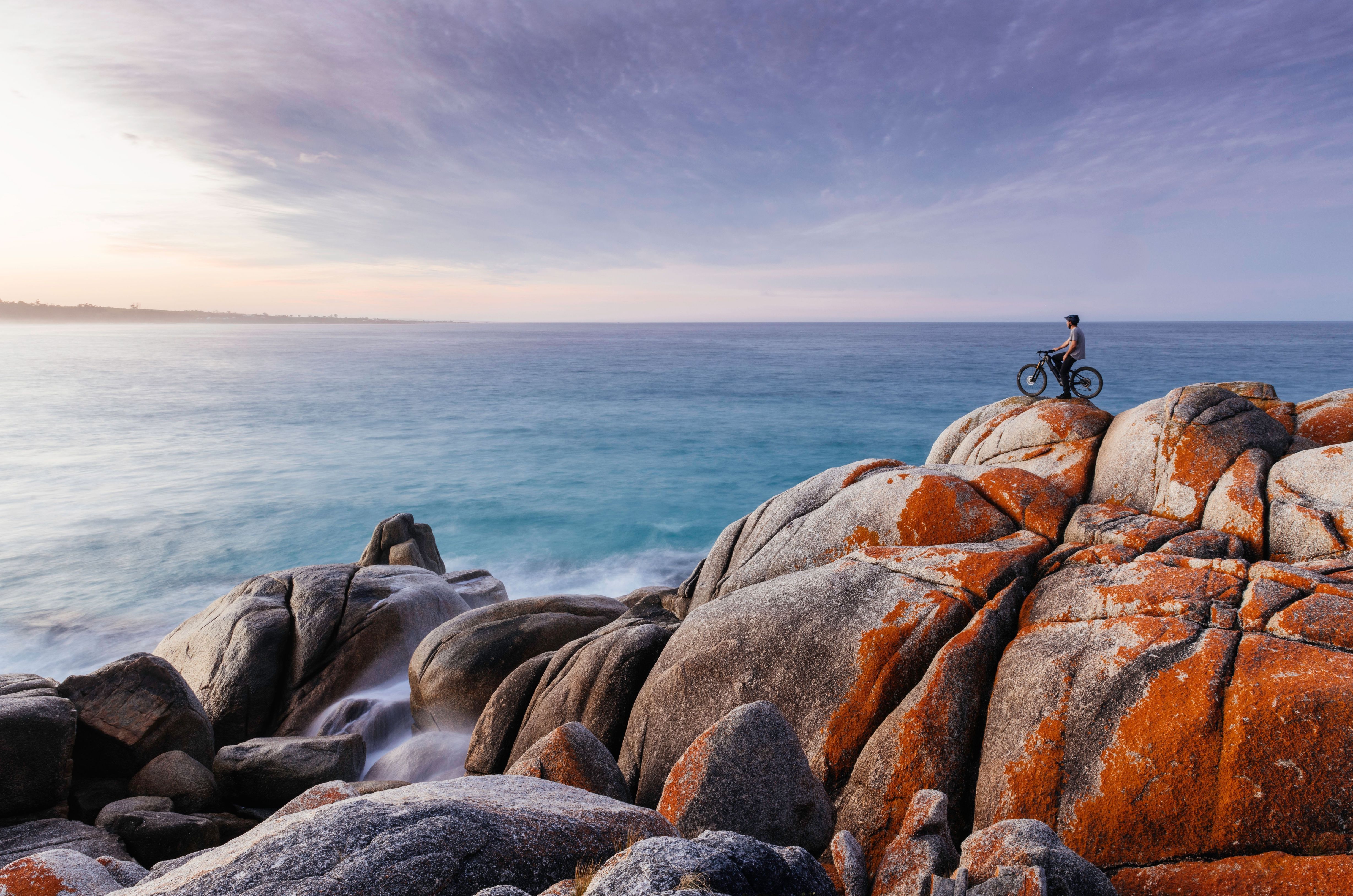 A cyclist standing on lichen covered granite boulders by the sea at larapuna / Bay of Fires area, Tasmania