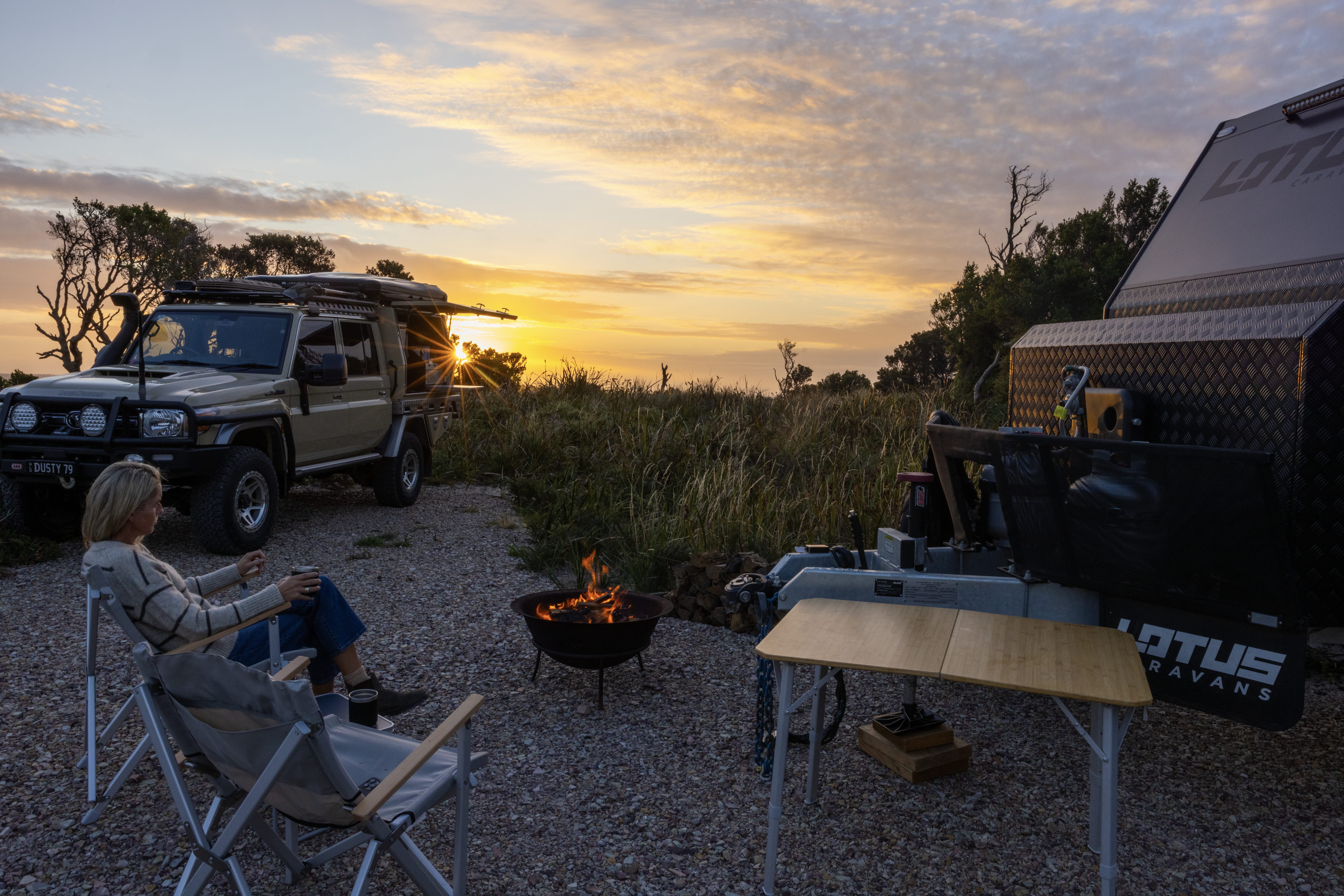 Person by a campfire at sunset beside a four-wheel drive and caravan at Cosy Corner South, Bay of Fires Conservation Area