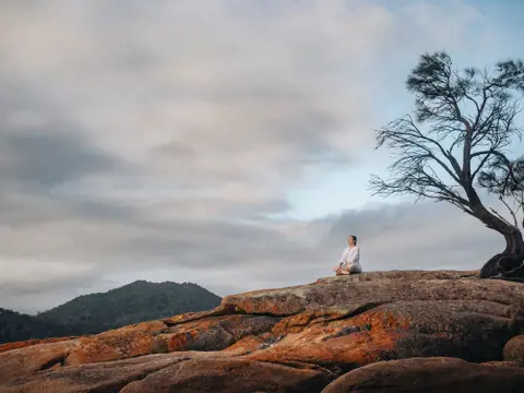 Person sitting cross-legged on the orange lichen-covered granite rocks at Honeymoon Bay, Freycinet National Park, Tasmania