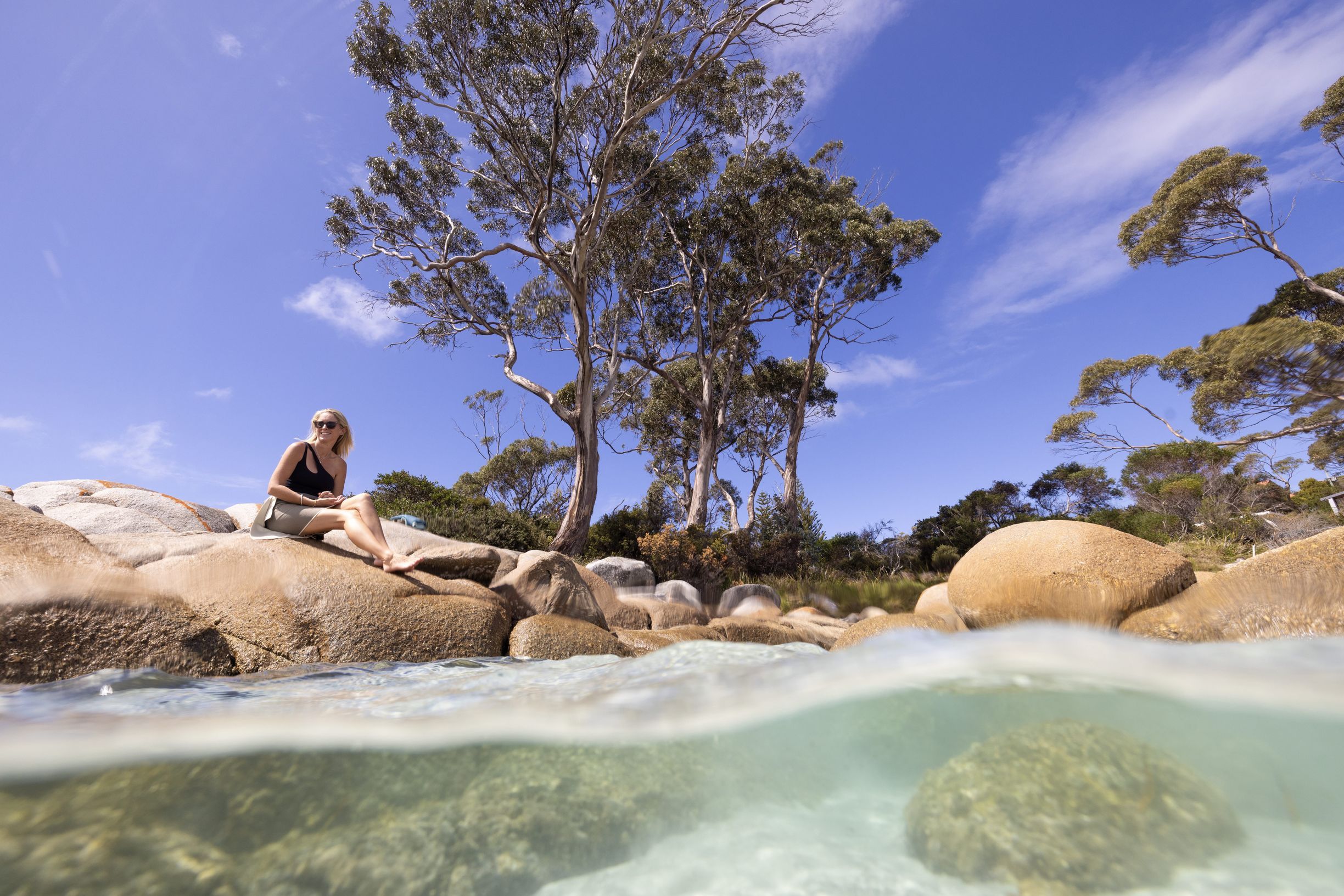 Person sitting on smooth boulders above clear shallow water at Waubs Bay Beach, Tasmania's east coast