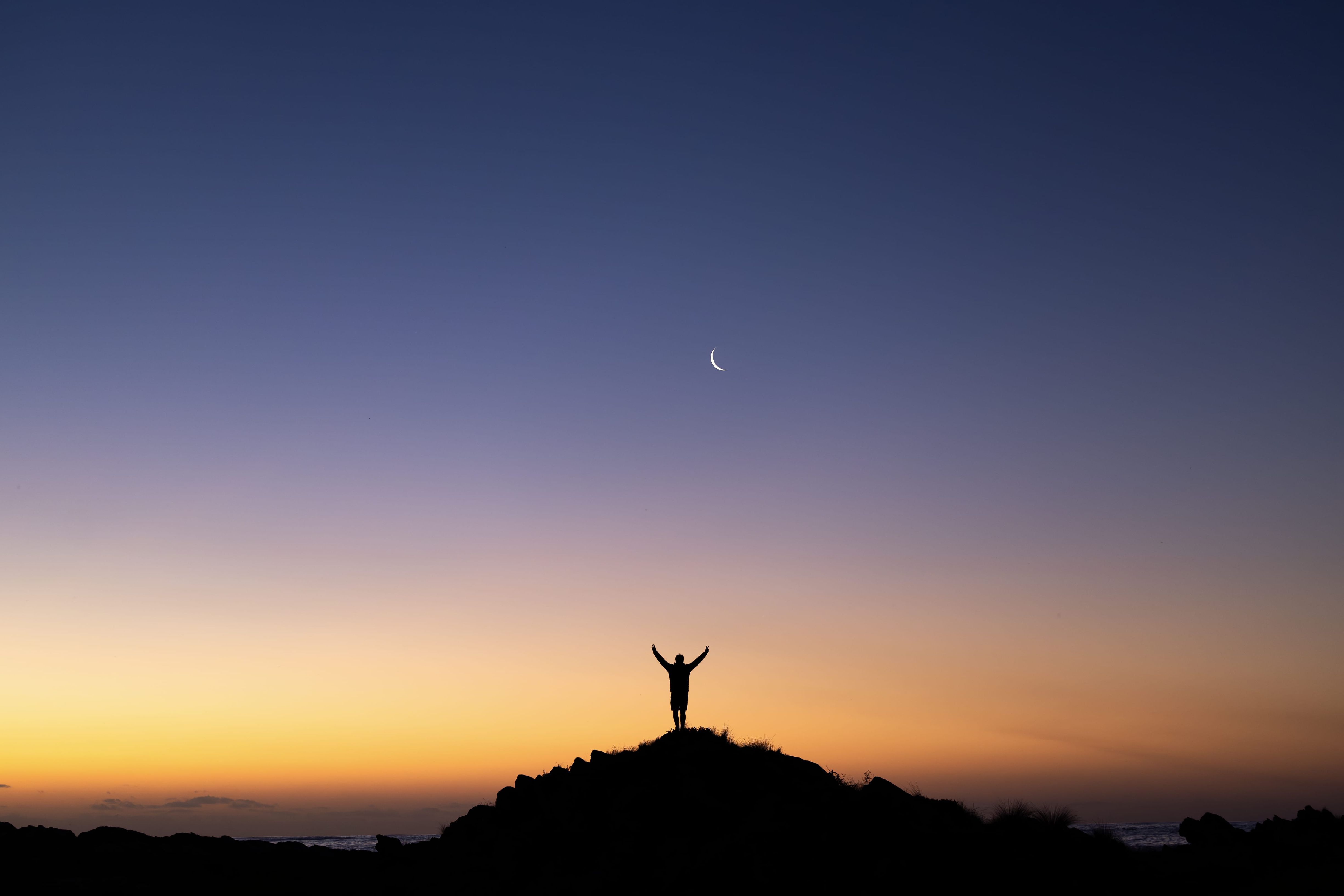 Silhouette of a person with arms raised on a rocky outcrop at sunset beneath a crescent moon, Arthur River, takayna / Tarkine