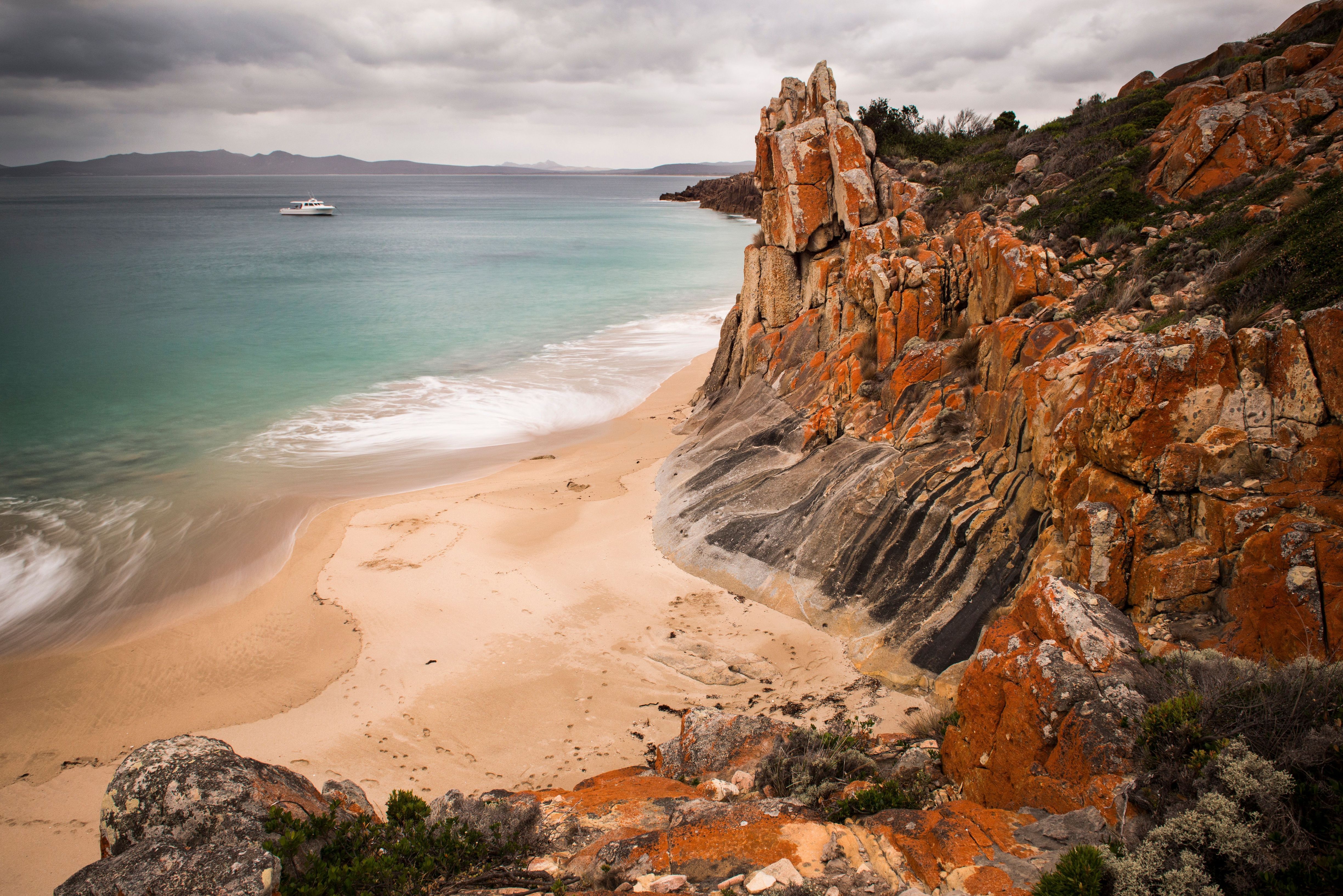 Boat anchored off a sandy beach with orange lichen-covered rocks under a cloudy sky north of Emita, Flinders Island