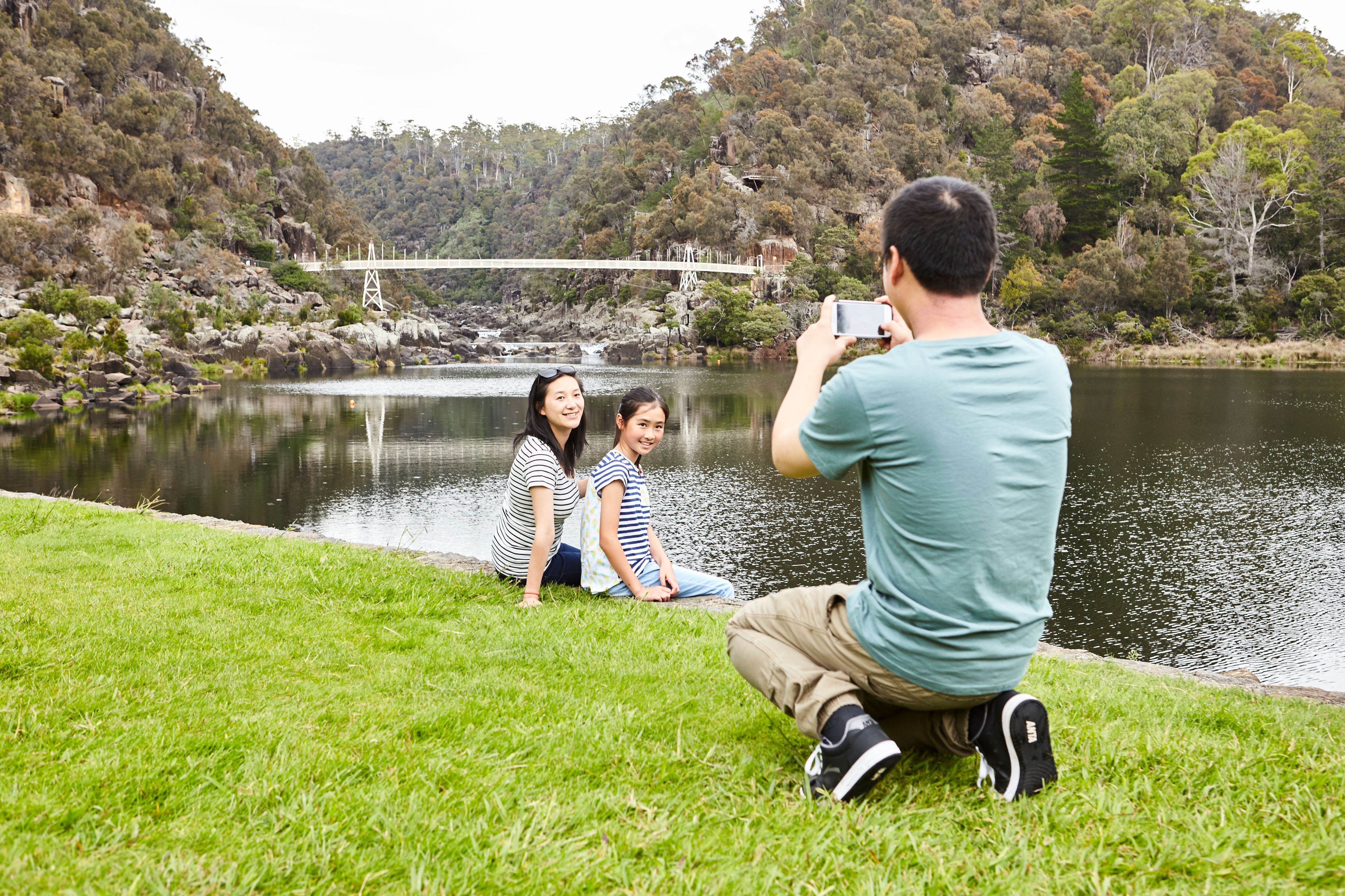 Person photographing two people by the water with the suspension bridge and cliffs behind at Cataract Gorge, Launceston