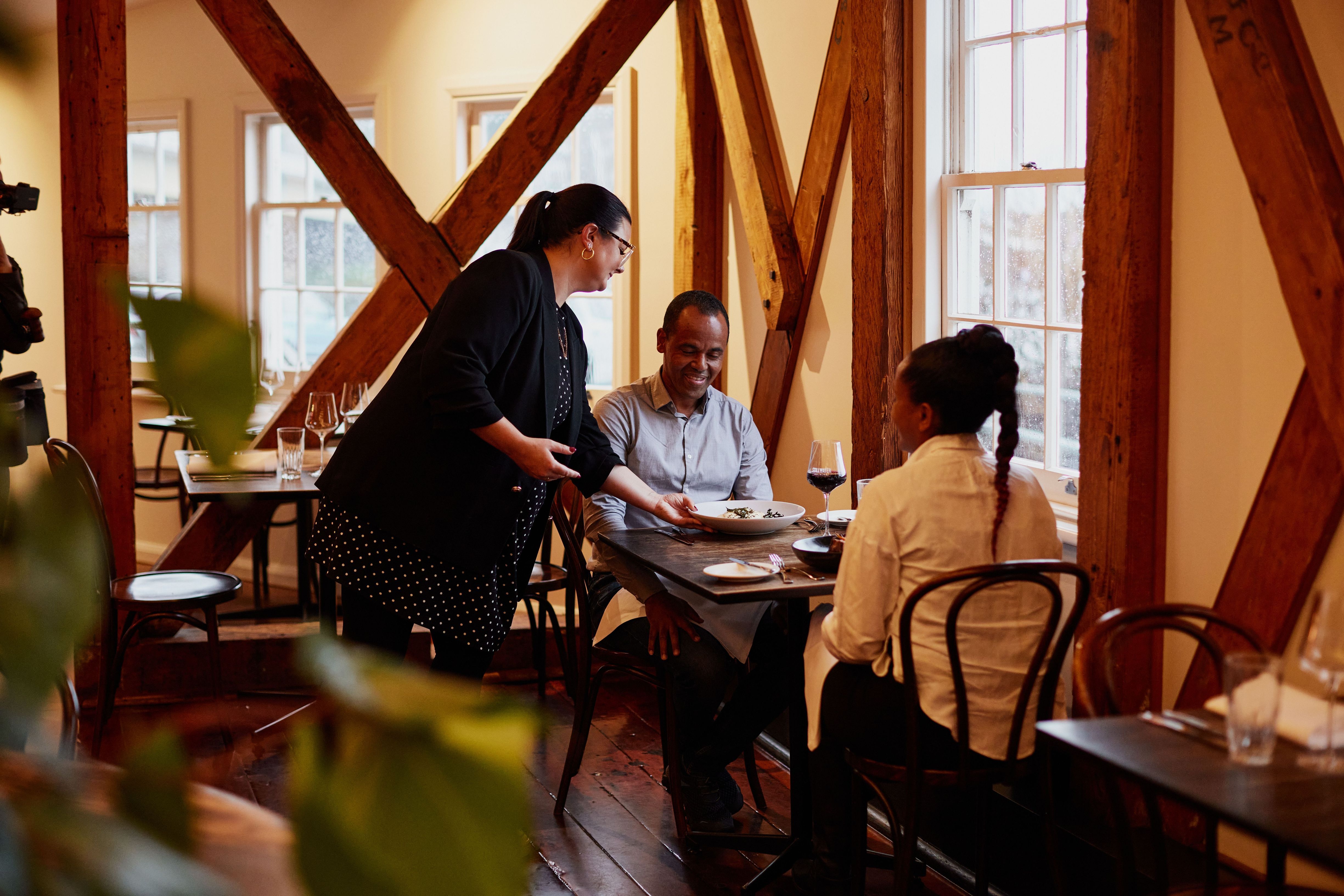 Server presenting a dish to two diners at a table by the window in the historic timber Stillwater restaurant, Launceston