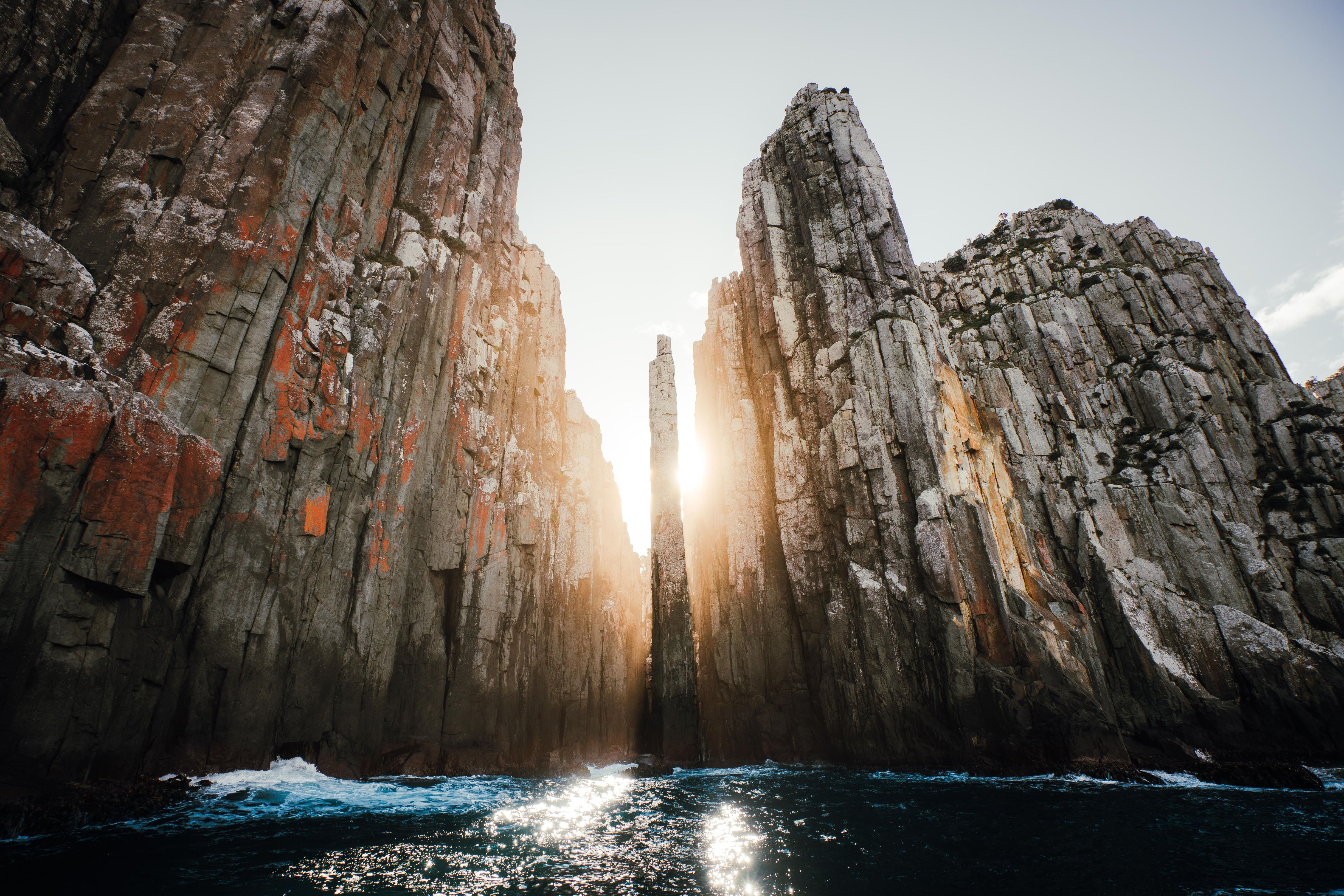 Sunlight streams between towering dolerite sea cliffs with a narrow rock pillar at The Candlestick, Cape Hauy, Turrakana / Tasman Peninsula