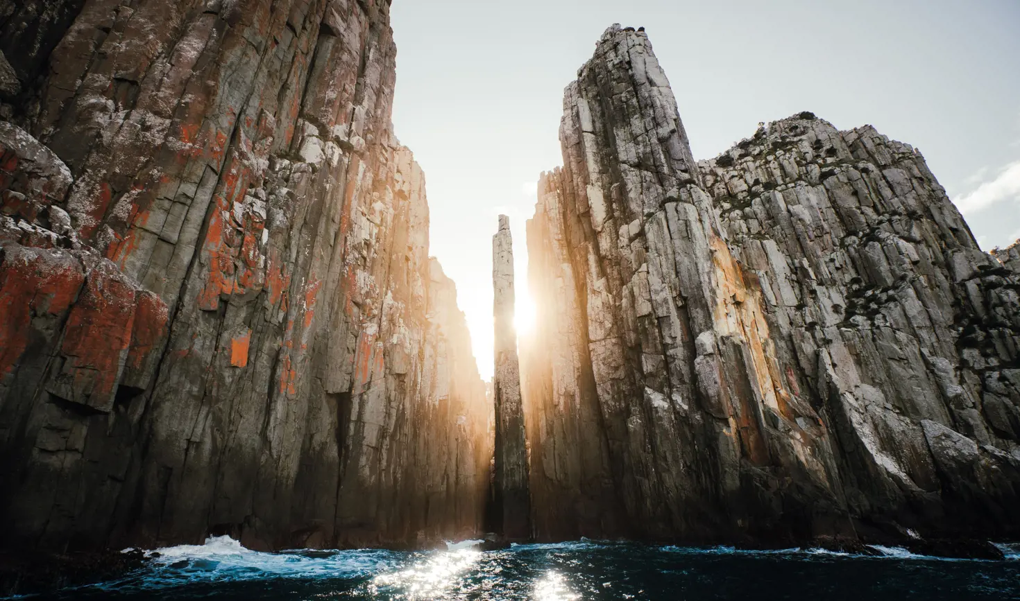 Sunlight streams between towering dolerite sea cliffs with a narrow rock pillar at The Candlestick, Cape Hauy, Turrakana / Tasman Peninsula