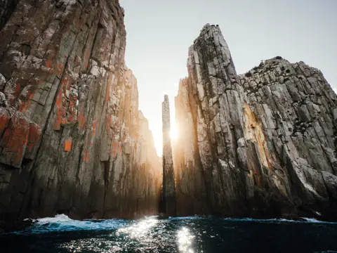 Sunlight streams between towering dolerite sea cliffs with a narrow rock pillar at The Candlestick, Cape Hauy, Turrakana / Tasman Peninsula