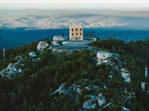 Aerial view of a sandstone tower building perched among granite boulders on a forested hilltop at The Keep in Goulds Country