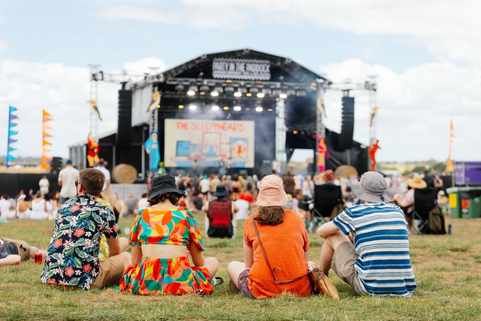 Four people sitting on the grass watching a band perform on the main stage at Party in the Paddock, Quercus Park, Carrick