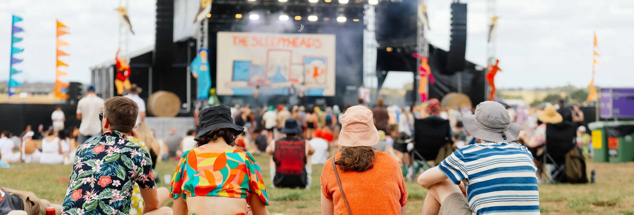Four people sitting on the grass watching a band perform on the main stage at Party in the Paddock, Quercus Park, Carrick