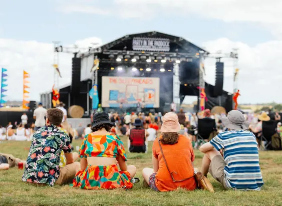 Four people sitting on the grass watching a band perform on the main stage at Party in the Paddock, Quercus Park, Carrick
