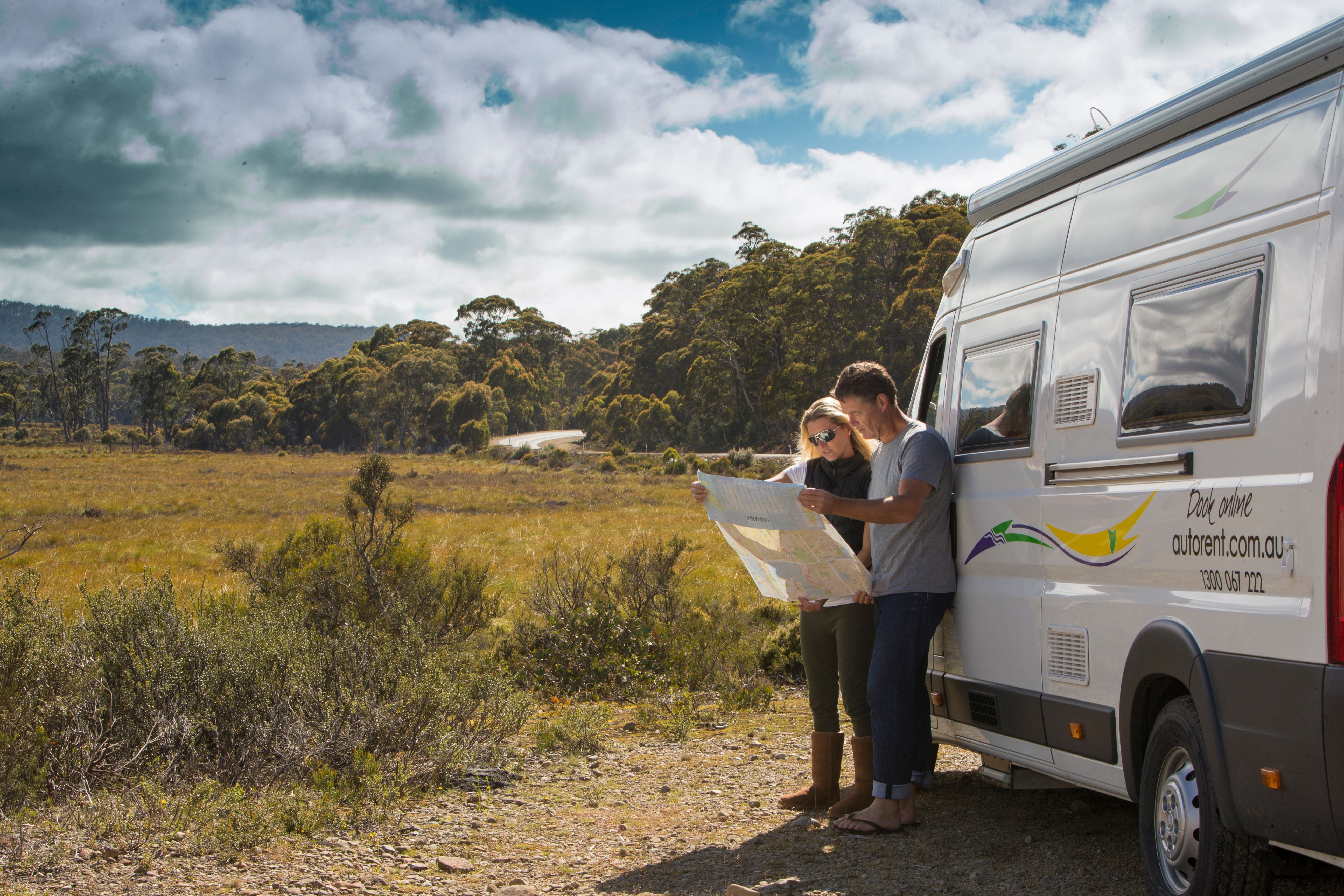 Two people read a map beside an Autorent campervan pulled over on a rural road with bushland and open plains, Huon Valley