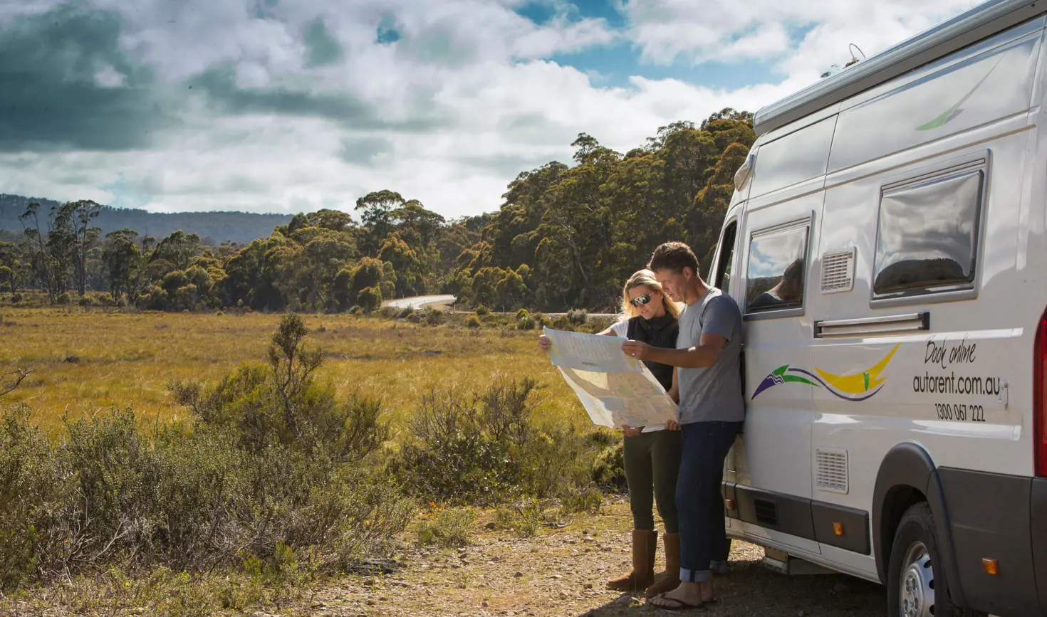 Two people read a map beside an Autorent campervan pulled over on a rural road with bushland and open plains, Huon Valley