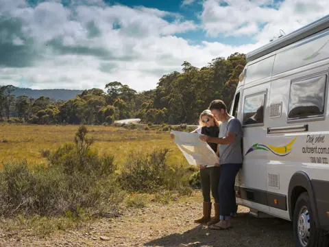 Two people read a map beside an Autorent campervan pulled over on a rural road with bushland and open plains, Huon Valley